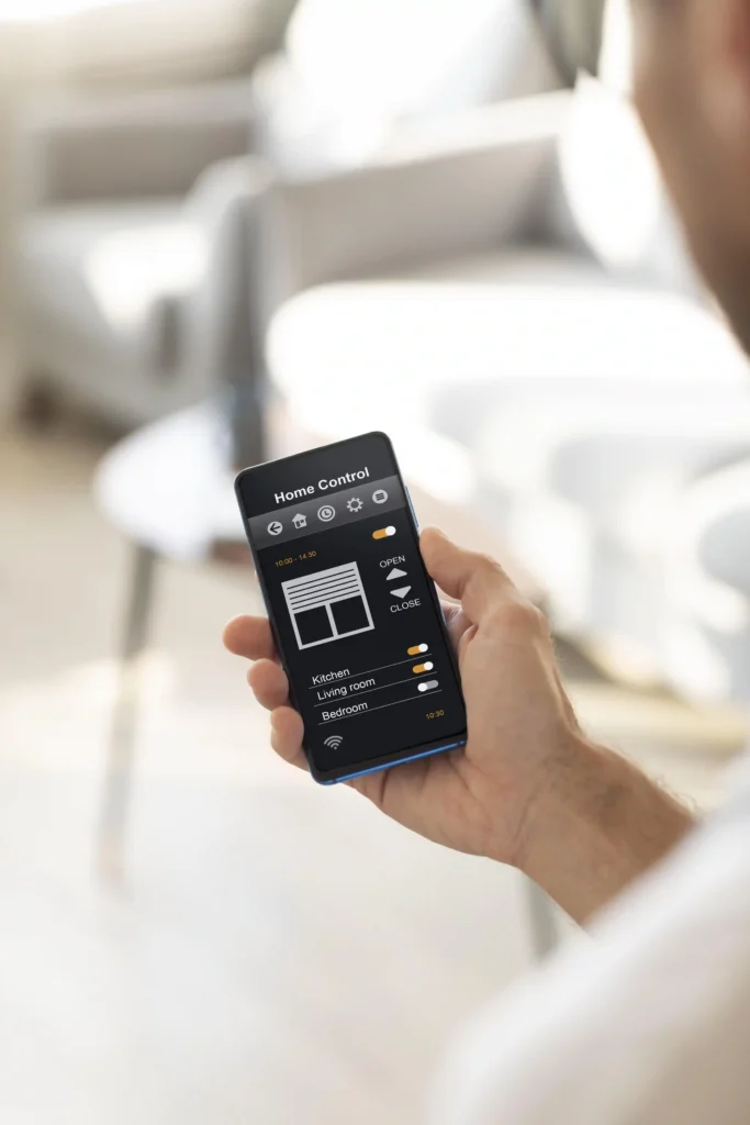 Close-up of a hand holding a smartphone showing a “Home Control” app with options to open blinds and toggle lights by room.