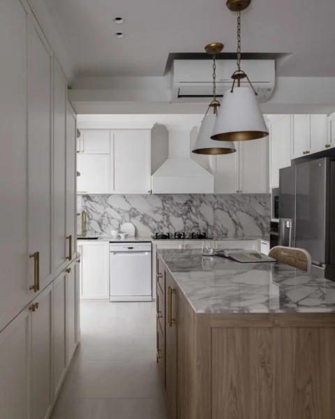 Classic white kitchen with marble backsplash, brass pendants, and a wood island topped with veined stone.