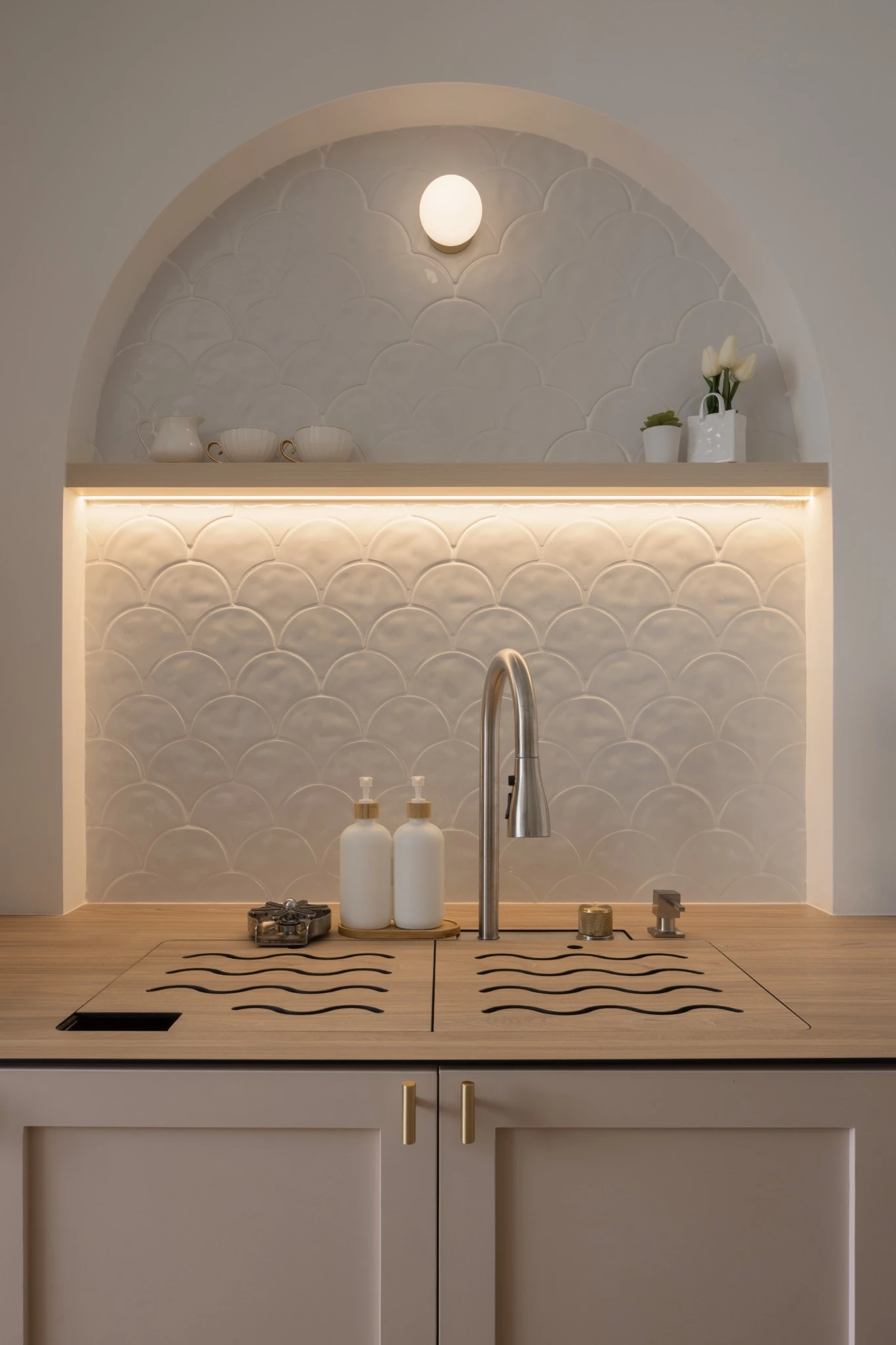 Kitchen sink with an arched niche, scallop-tile backsplash, under-shelf lighting, and simple countertop accessories.