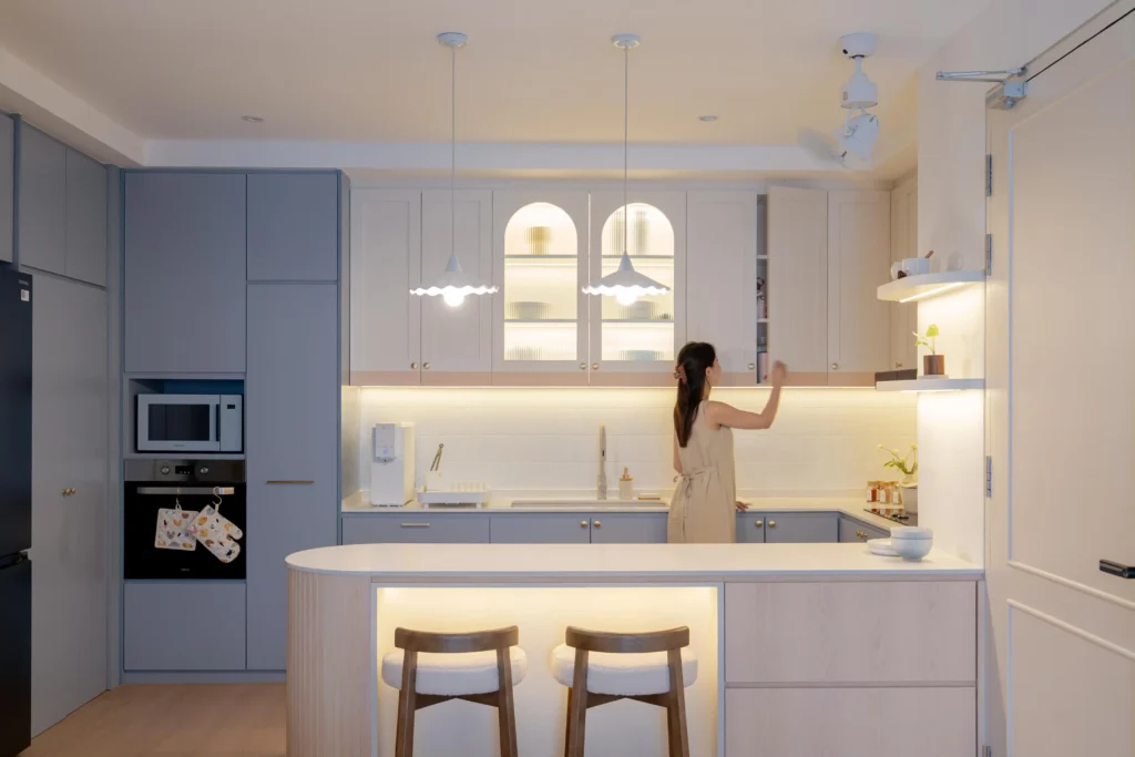 Modern kitchen with blue cabinets, arched glass display units, a fluted island, and pendant lights; a woman reaching for a shelf.